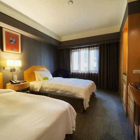 Ambassador Hotel Kaohsiung Exterior foto The photo shows a hotel room with two beds, each made up with white linens. The walls are painted in dark colors, while the bedding and furniture are lighter in tone. There is a nightstand between the beds with a lamp and telephone on it. A window wi