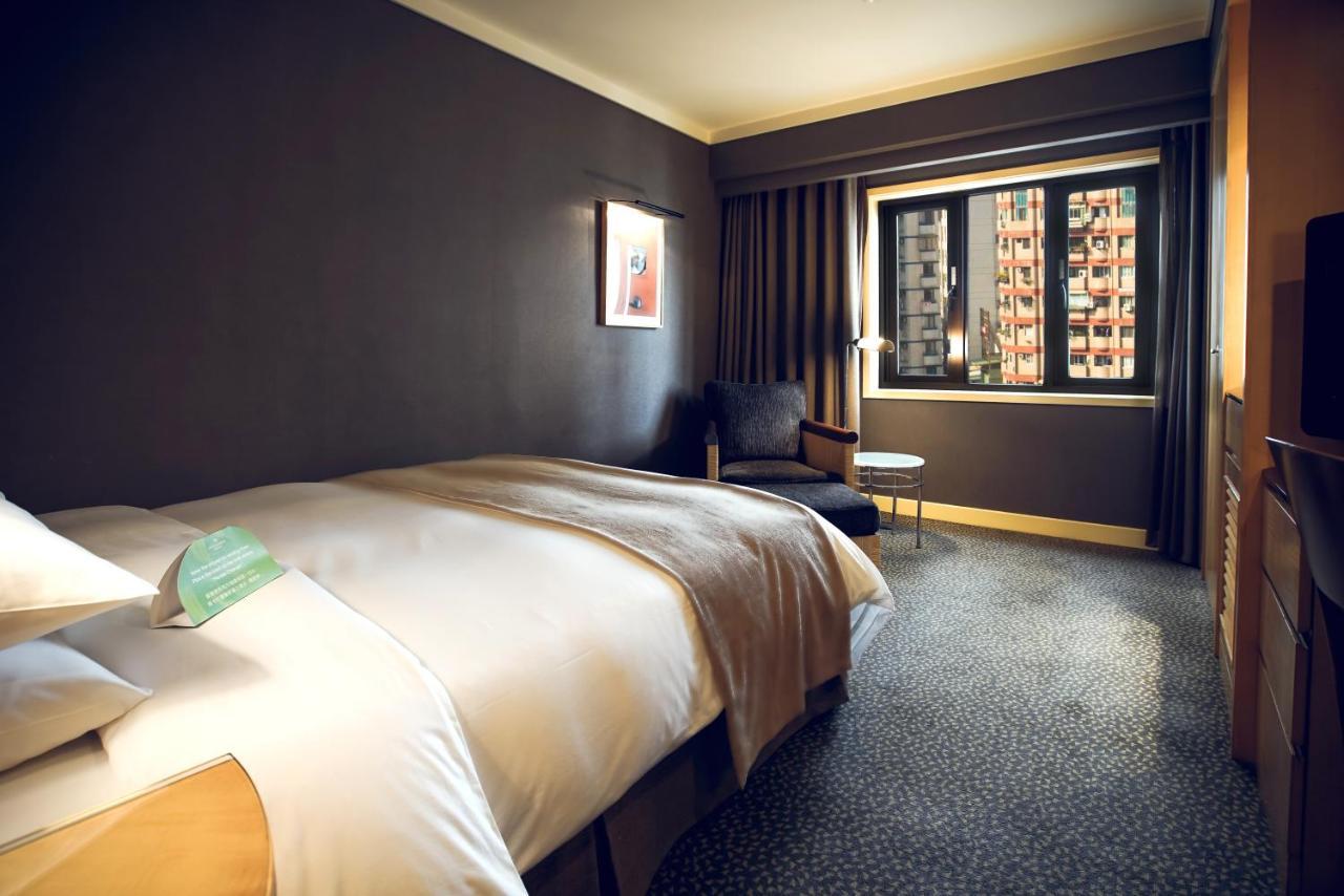 Ambassador Hotel Kaohsiung Exterior foto The photo shows a hotel room featuring a large bed with a light-colored duvet. The walls are painted in a dark hue, creating a cozy atmosphere. There is a chair and a small side table near the window, which is covered by curtains. Through the window,