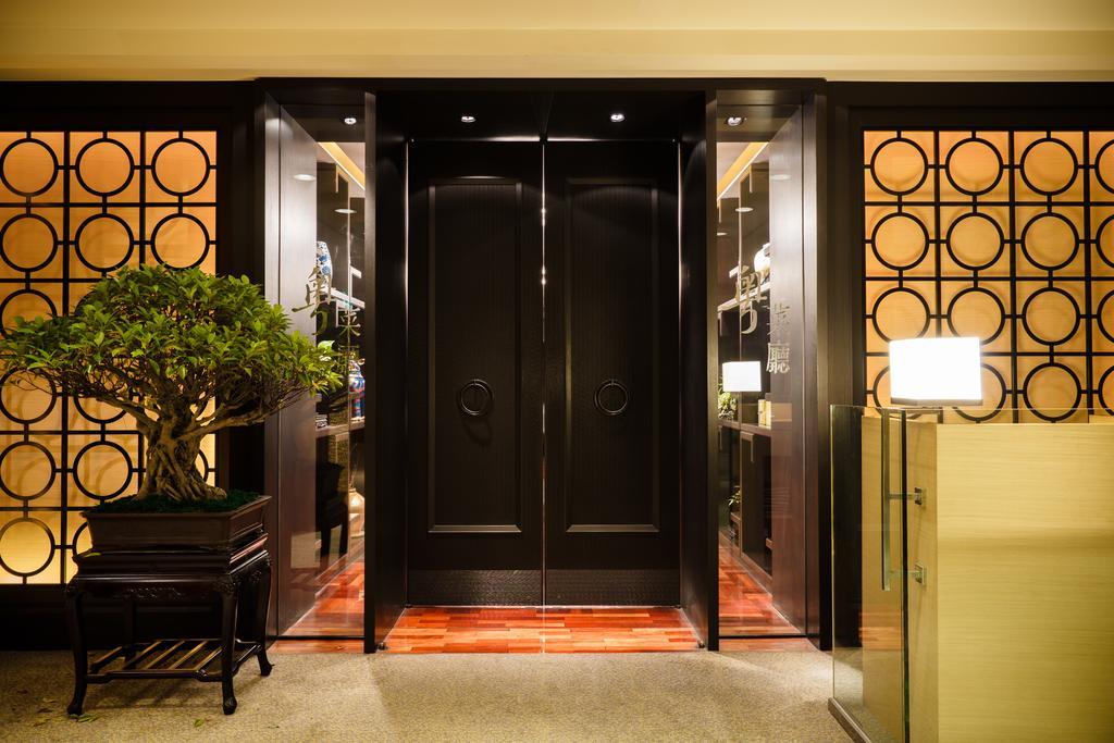 Ambassador Hotel Kaohsiung Exterior foto The photo shows an elegant entrance area featuring a set of large black double doors, which appear to be a main entryway. The walls surrounding the doors have a decorative design with circular patterns. On one side, there is a small tree in a planter