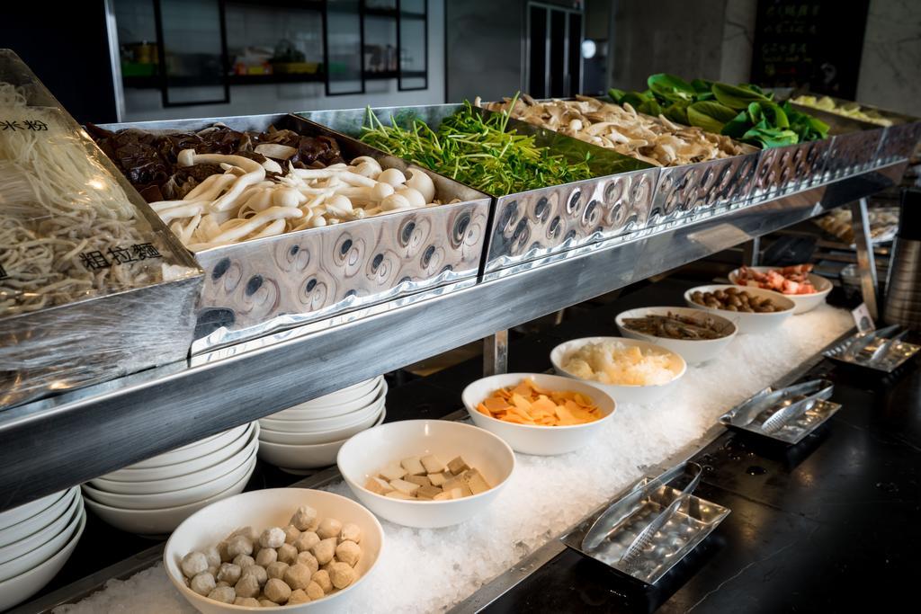 Ambassador Hotel Kaohsiung Exterior foto The photo shows a buffet or display of various food items, likely from an Asian cuisine setting. There are several metal trays filled with a variety of fresh vegetables, including greens and mushrooms. Below these trays, there are white bowls contain