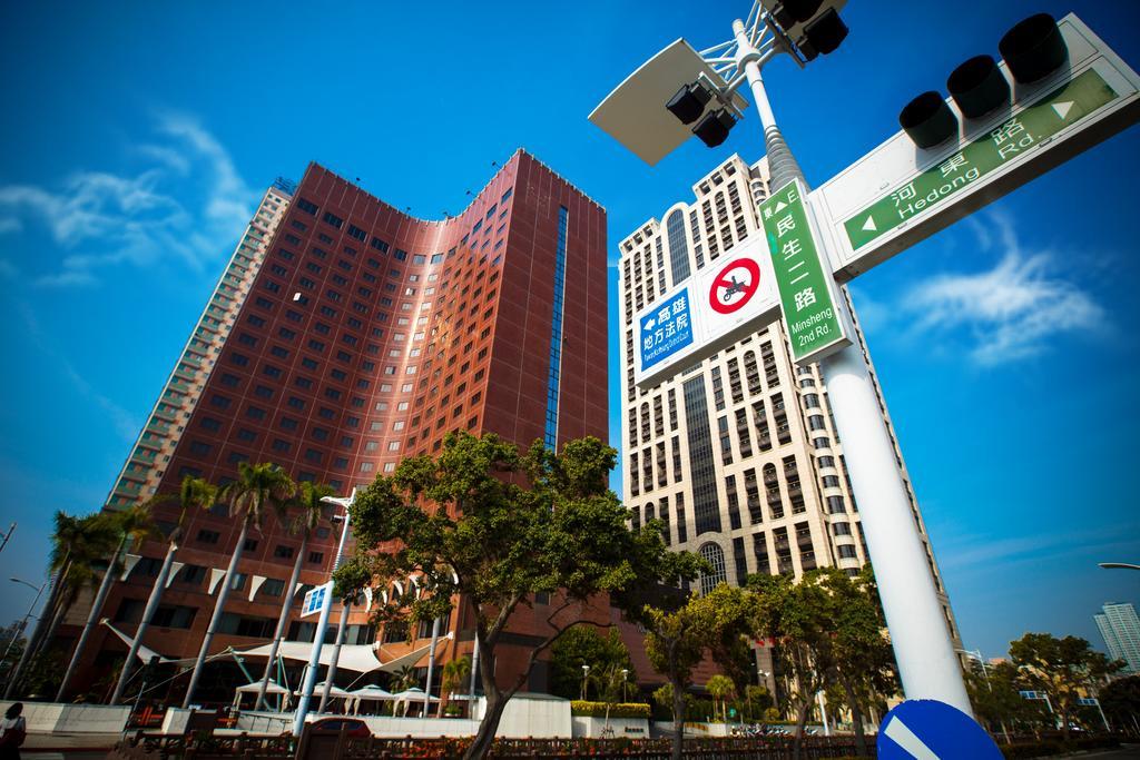 Ambassador Hotel Kaohsiung Exterior foto The photo shows a cityscape with tall buildings, including one red-brick structure with a unique architectural design. The sky is clear and blue, suggesting a bright and sunny day. In the foreground, there are traffic signs, including a direction sig