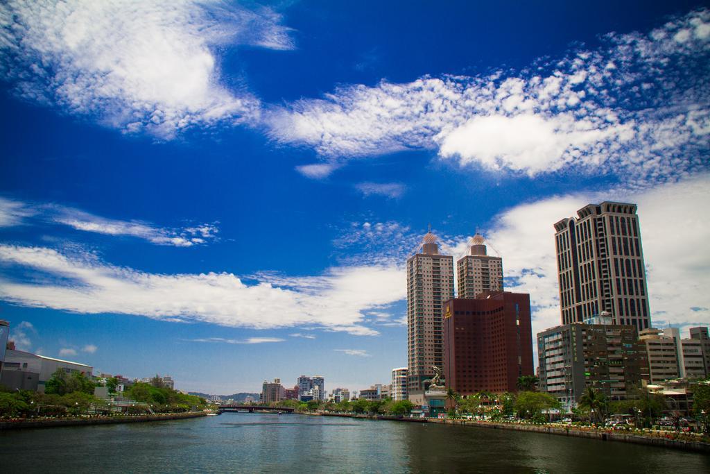 Ambassador Hotel Kaohsiung Exterior foto The photo showcases a scenic view of a river flanked by modern skyscrapers. The buildings have a mix of architectural styles, including tall, glass-fronted structures and a reddish-brown building. In the background, a clear blue sky features fluffy w