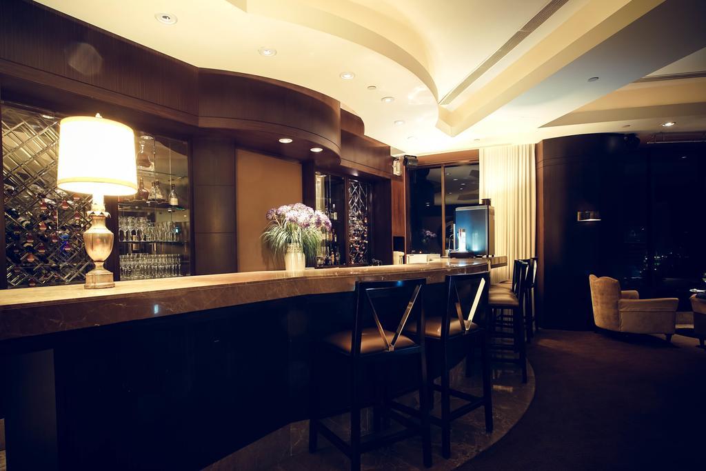 Ambassador Hotel Kaohsiung Exterior foto The photo shows a modern and stylish bar area. The bar is made of a smooth stone surface and features a backlit shelf displaying various bottles of liquor. There is a lamp on the bar providing warm lighting, and a vase with flowers adds a decorative 