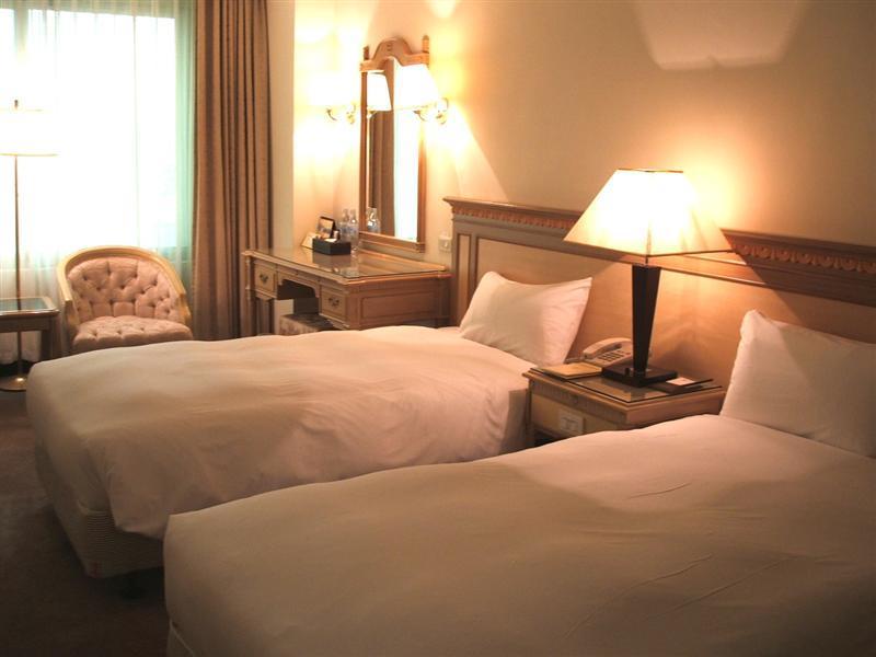 Ambassador Hotel Kaohsiung Exterior foto The photo shows a cozy hotel room with two single beds, each covered with white bedding. There is a lamp on a nightstand between the beds. A wooden dresser with a mirror is against the wall, and a comfortable armchair is placed nearby. Soft lighting 