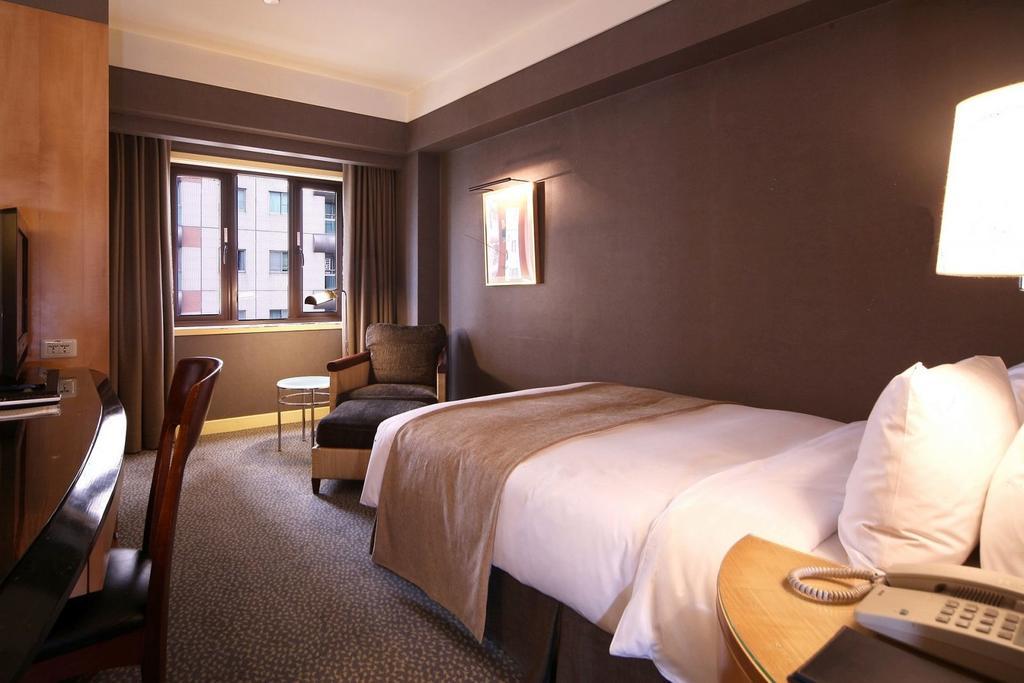 Ambassador Hotel Kaohsiung Exterior foto The photo shows a hotel room featuring a neatly made single bed with a light-colored blanket. There is a comfortable chair in the corner alongside a small round table. A large window allows natural light to enter the room, and the walls are painted i