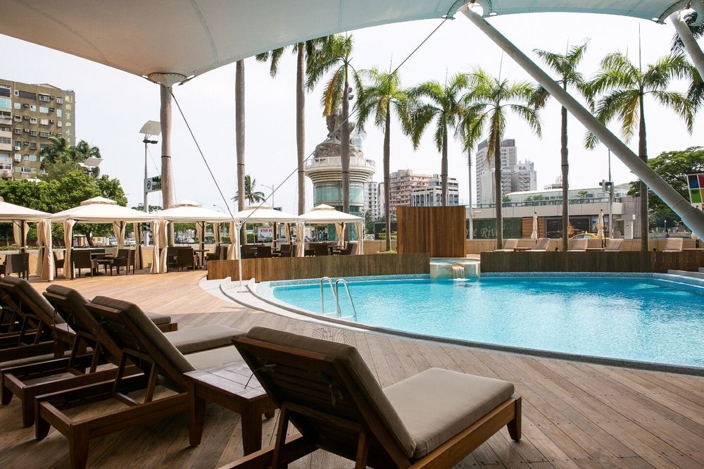 Ambassador Hotel Kaohsiung Exterior foto The photo shows a resort-style pool area. In the foreground, there are several wooden lounge chairs arranged around a sparkling blue pool. The pool has a gentle slope and is surrounded by wooden decking. Behind the pool, tall palm trees add to the tr