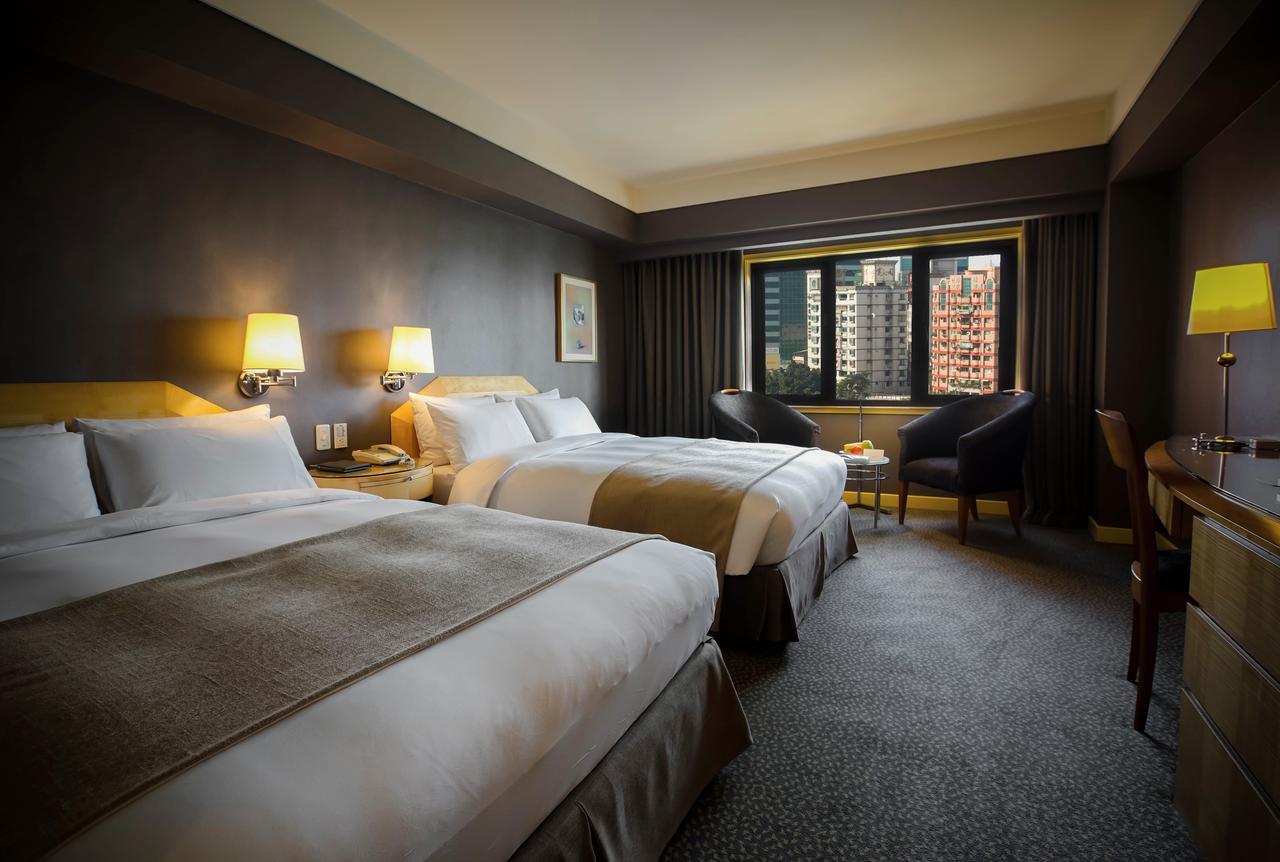 Ambassador Hotel Kaohsiung Exterior foto The photograph shows a hotel room featuring two double beds with white linens and light brown accents. The walls are painted a dark color, and there are bedside lamps beside each bed. A window offers a view of the outside, and there are two chairs po