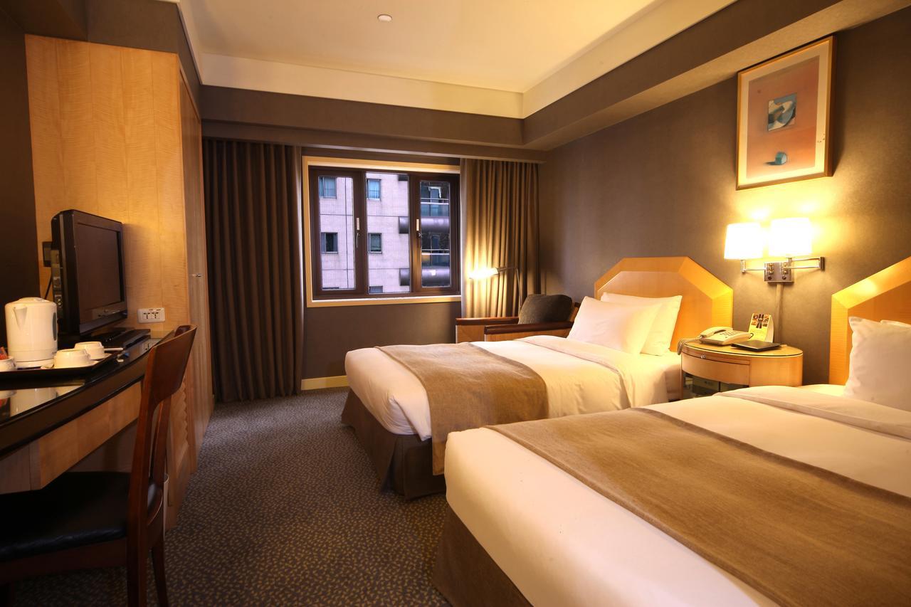 Ambassador Hotel Kaohsiung Exterior foto The photo shows a hotel room featuring two twin beds with neutral-colored bed linens. The room has a modern design, with warm lighting and wooden furnishings. There's a desk and chair positioned against one wall, along with a television. A large wind