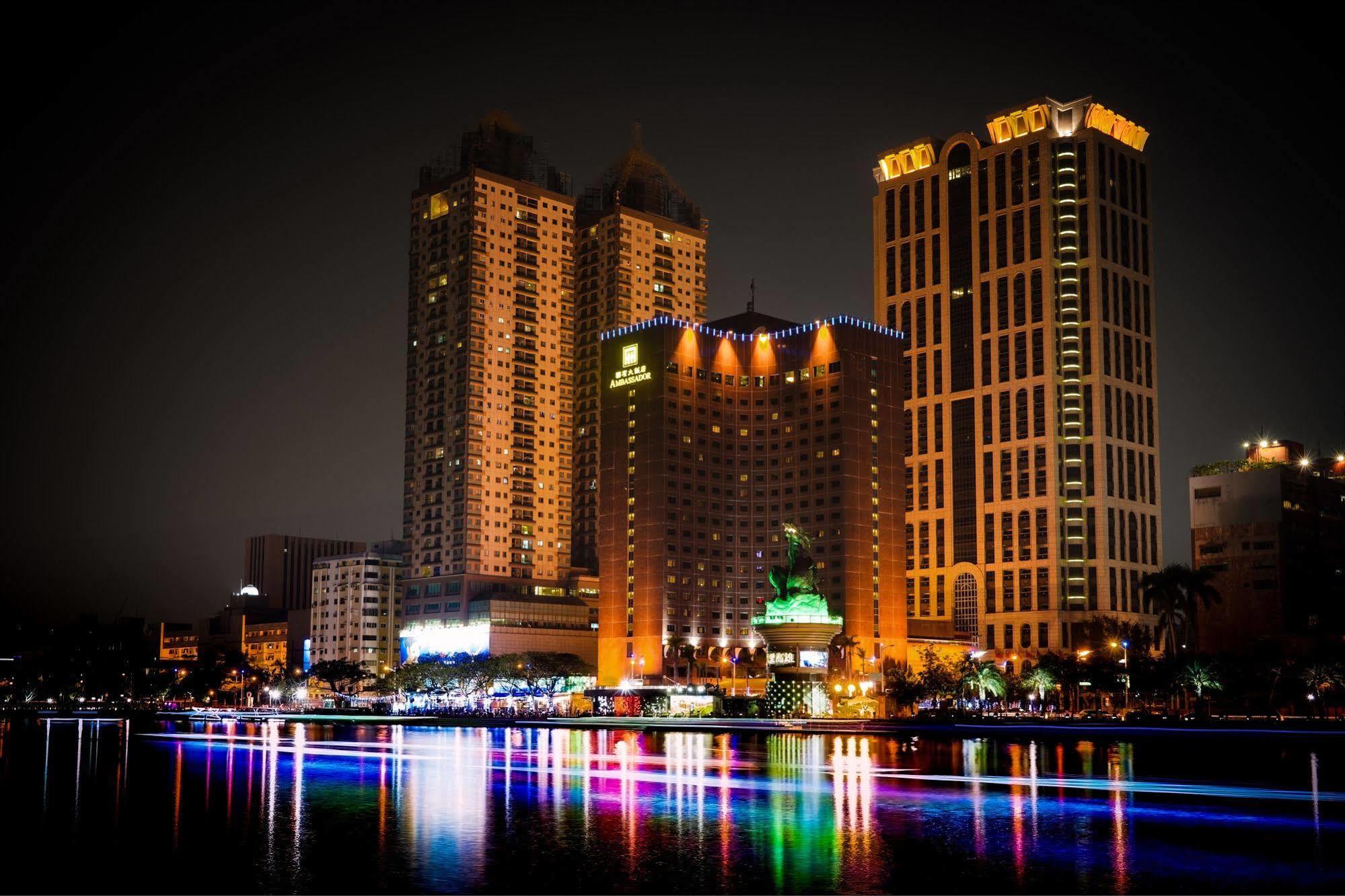 Ambassador Hotel Kaohsiung Exterior foto The photo depicts a vibrant nighttime cityscape featuring several tall buildings illuminated by colorful lights. Prominent structures include a large hotel and other high-rise buildings with illuminated facades. In front of these buildings, there app
