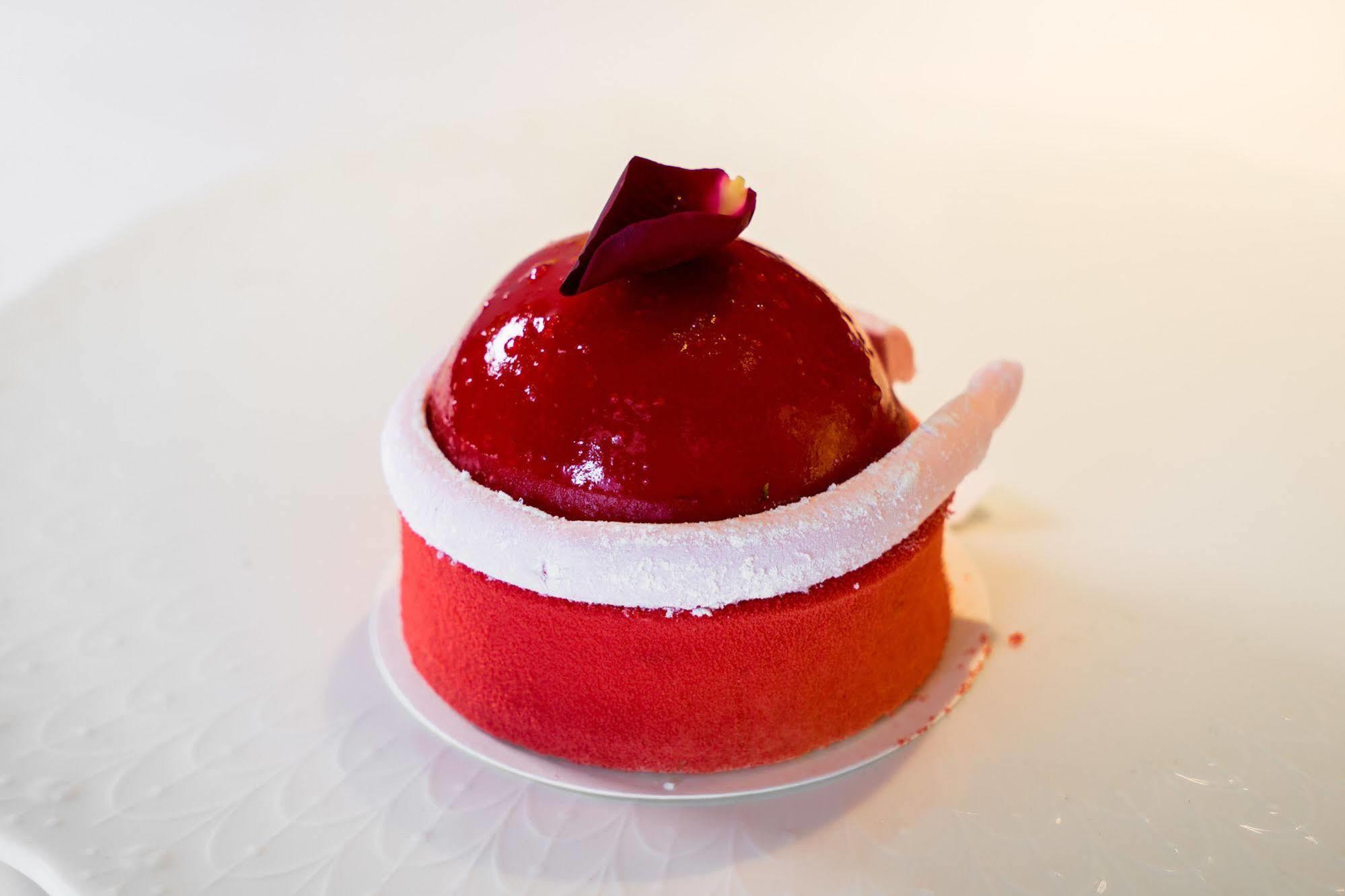 Ambassador Hotel Kaohsiung Exterior foto The photo shows a beautifully crafted dessert that appears to be a mousse or cake. It has a glossy red coating, shaped like a dome on top, with a small decorative rose petal placed on it. The base of the dessert features a gradient of colors, transit