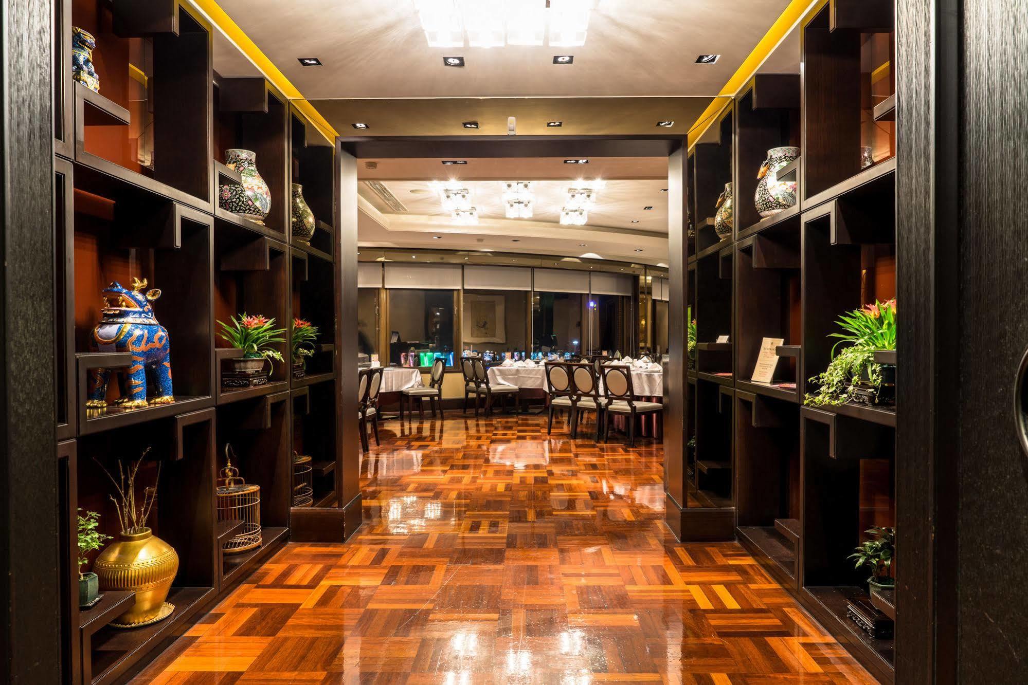 Ambassador Hotel Kaohsiung Exterior foto The photo shows the interior of a restaurant or dining area. It features a corridor lined with dark wooden shelves displaying decorative items and plants. The flooring is made of polished wood, which reflects the warm lighting from the ceiling fixtur