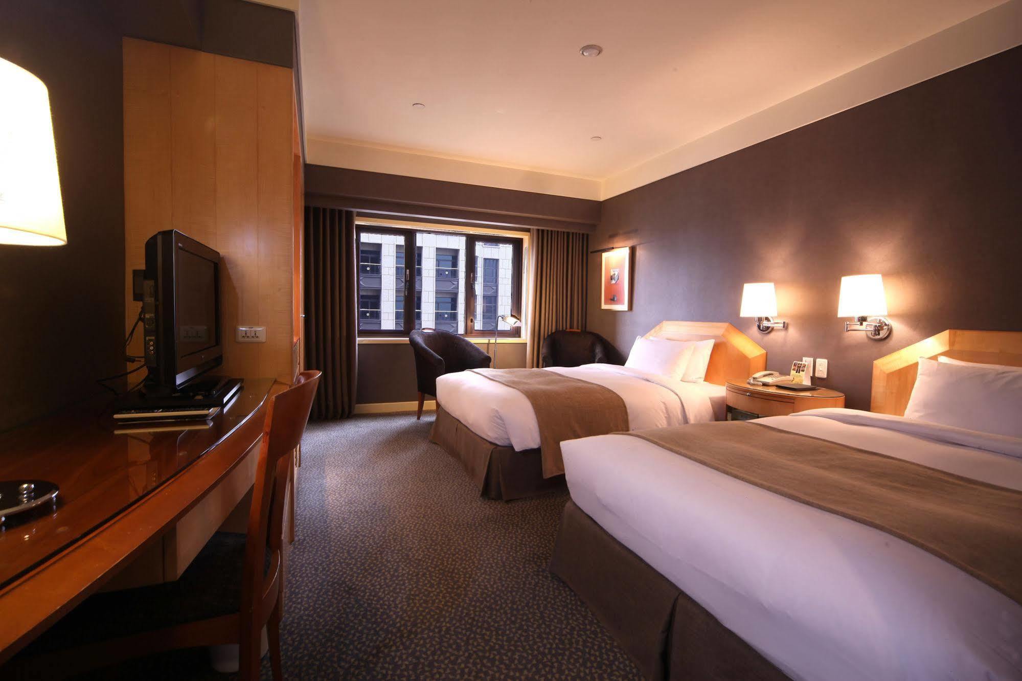 Ambassador Hotel Kaohsiung Exterior foto The photo shows a hotel room featuring two twin beds, each with light-colored bedding. There are bedside lamps on each side of the beds. A wooden desk with a chair is positioned next to a wall-mounted television. The room has a large window with shee