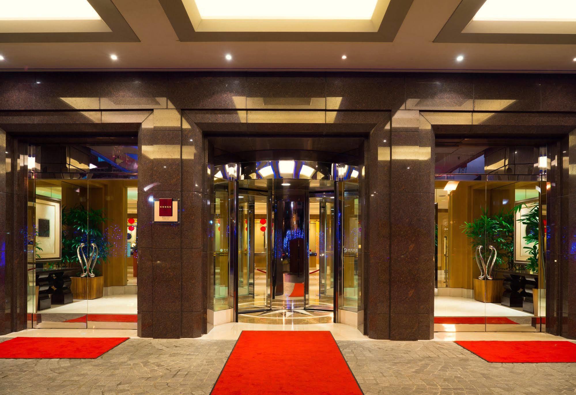 Ambassador Hotel Kaohsiung Exterior foto The photo shows the entrance of a modern hotel or building. It features large glass doors in a sleek frame, creating an inviting entrance. The floor is covered with a plush red carpet leading up to the doors. Inside, there are decorative plants on ei