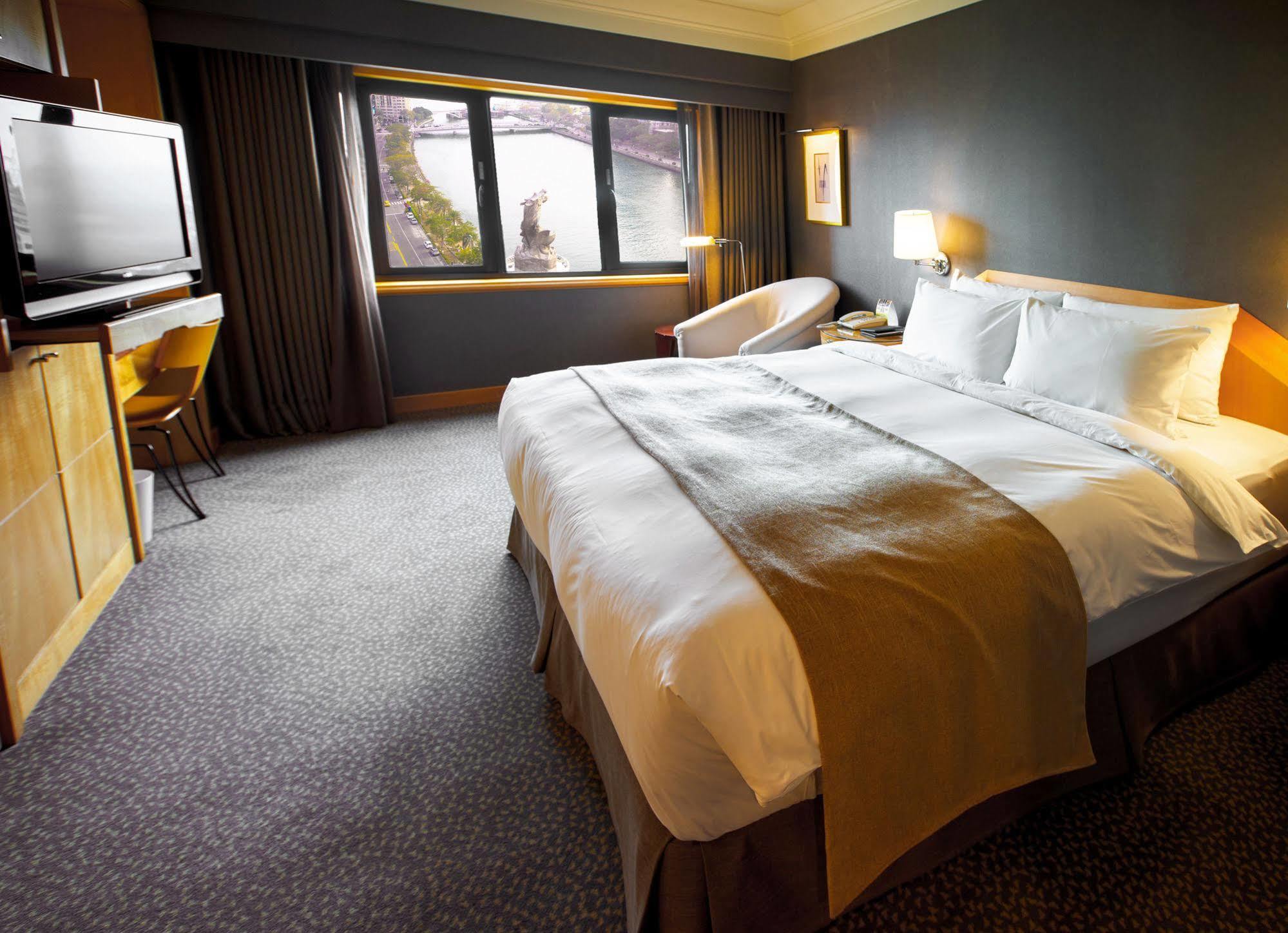 Ambassador Hotel Kaohsiung Exterior foto The photo depicts a modern hotel room featuring a spacious layout. In the center, there is a large bed with crisp white linens and a decorative brown throw blanket. To the side, there is a comfortable armchair and a small table. A television is mount