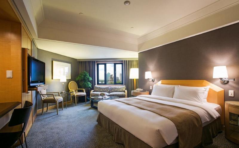 Ambassador Hotel Kaohsiung Exterior foto The photo shows a hotel room that features a large bed with white linens and a light brown throw blanket. There are two bedside lamps on either side of the bed. In the corner, there is a cozy seating area with a patterned sofa and two chairs. A telev