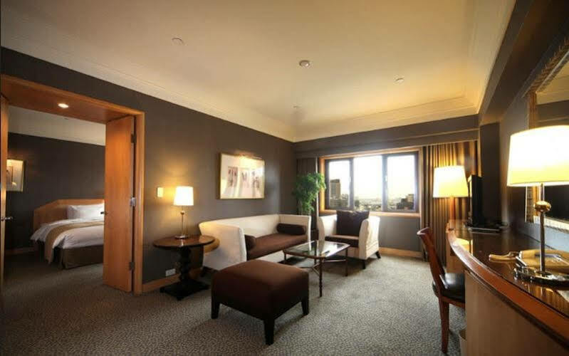 Ambassador Hotel Kaohsiung Exterior foto The photo shows a stylish hotel room with modern décor. There is a comfortable seating area featuring a white sofa and brown ottoman, along with a glass coffee table. A side table with a lamp is positioned next to the sofa. The room has a large windo