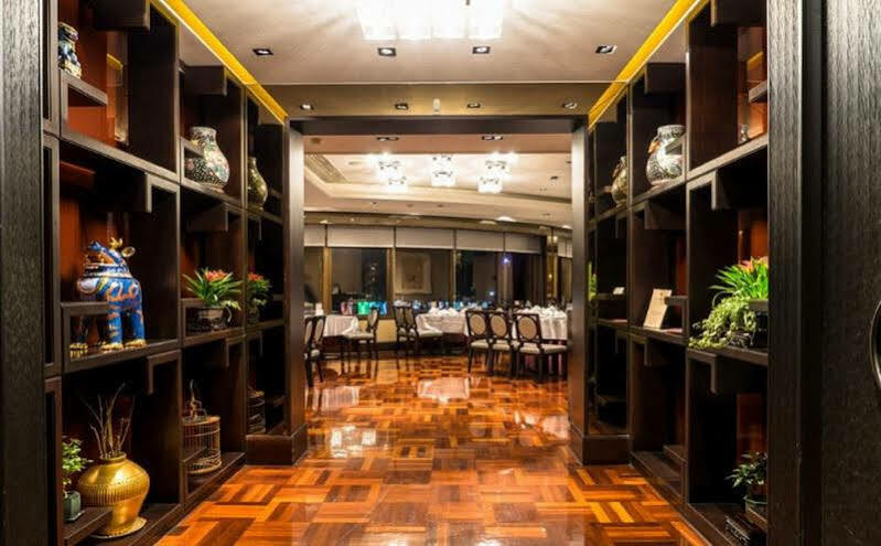 Ambassador Hotel Kaohsiung Exterior foto The photo shows an elegant restaurant interior. In the foreground, there are dark wooden shelves filled with decorative items, such as vases and small sculptures, along with plants. The floor is made of polished wood, featuring a geometric pattern. I